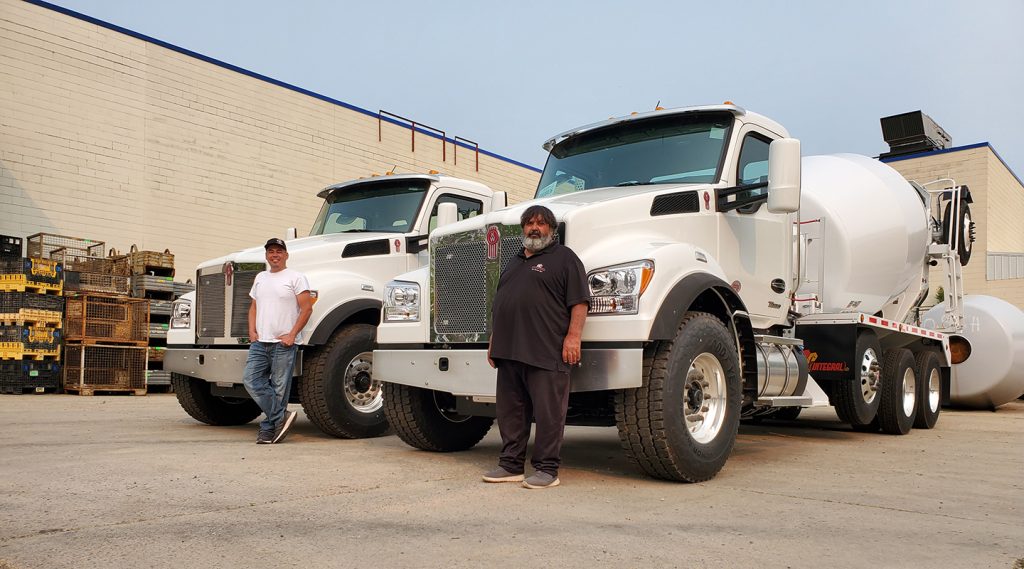 A pair of men standing in front of a pair of INTEGRALdx concrete redi-mix trucks with booster axles at the rear. Each truck has a white Kenworth chassis.