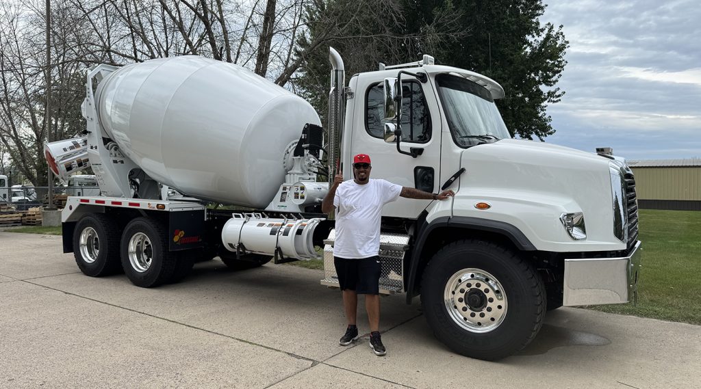 A smiling man standing in front of a white INTEGRALdx concrete redi-mix truck in the conventional style. The man has one thumb up. The truck's chassis was designed by Freigtliner.