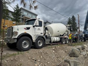 The front left view of an INTEGRALdx concrete redi-mix booster truck with a drum fill line of 14 cubic yards of concrete. It is parked on a rocky, inclined roadway. The driver-side door of the Peterbilt chassis has a decal for M&K Ready Mix Inc. Two men with construction vests stand near the ladder in the rear.