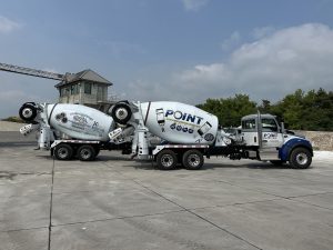 The right side view of two INTEGRALdx concrete redi-mix booster trucks made for Point Ready Mix. They feature Kenworth chassis colored gray, white, and navy blue. The passenger side doors read Point Ready Mix. Additionally, the back left truck has a drum with the text: Proud Sponsors of Gigi's Playhouse, trademarked. Down Syndrome Achievement Centers. Educate. Inspire. Believe. McHentry County, Illinois. 20 years of believing. Gigi's Playhouse. Please join us in sponsoring, donating, and volunteering. A QR code can be found on the side of the truck. On the front truck, the drum reads: Point. Customers love our mobile app! Real-time view of all jobs. See your trucks on a map. Receive alerts. Order calendar. Two animated construction workers with hard hats and vests hold two cell phones. The one on the right states, 'With Point I get an alert on my phone when my truck is 15 minutes away!' A QR code is found to the worker's left.