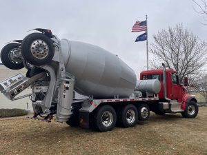 A rear right view of an INTEGRALdx concrete redi-mix booster truck on the front lawn of the SpecSys facility in New Ulm. This truck has a red Peterbilt chassis and a gray drum.