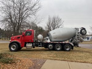 The left side view of an INTEGRALdx concrete redi-mix booster truck on the front lawn of the SpecSys facility in New Ulm. This truck has a red Peterbilt chassis and a gray drum.