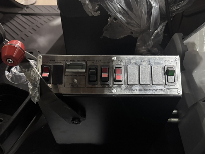 The in-cab cable controls of an INTEGRALdx concrete redi-mix truck. This particular model features a red lever. There is also a drum counter and a temperature light (green means normal; red means high). Furthermore, the control box has toggle switches for the following features: starting or stopping the drum, charging the drum, lifting up the chute, locking the chute, activating or deactivating the hopper, and toggling the work light on or off.
