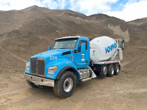 The front left view of an INTEGRALdx concrete redi-mix booster truck parked at a construction site. The driver-side door of the light blue Kenworth chassis has a decal for Ioppolo Concrete Corp. The white water tank and drum have blue Ioppolo logos along the side.